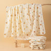 A white soft cotton gauze baby swaddle blanket draped over a wooden rod, with folded blankets nearby.