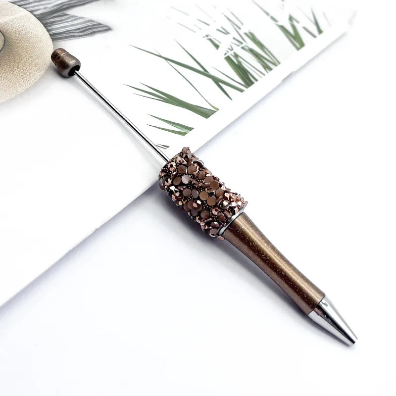 A close-up of a gold-colored ballpoint pen with a sequin-covered tip resting on a piece of paper featuring green grass design