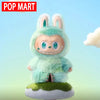 A light green Labubu plush doll with large ears and a smiling expression sitting on a mossy base.