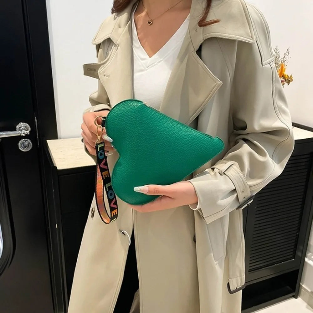 A hand holds a green heart-shaped PU coin purse with a leather strap, displayed against a light beige trench coat.