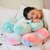 A person is resting their head on a large, fluffy, light blue and pink sunflower plush pillow in a bright bedroom setting.