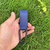 A hand holds a blue PU leather stroller hook with a black loop against a green grassy background.