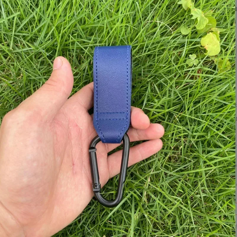A hand holds a blue PU leather stroller hook with a black loop against a green grassy background.