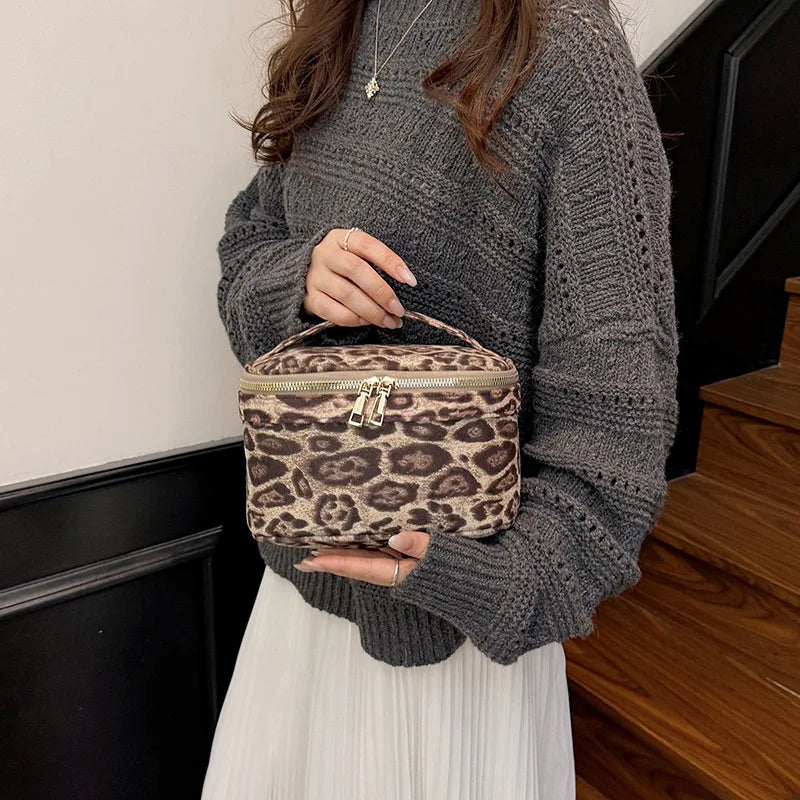 A woman wearing a gray sweater holds a brown leopard print cosmetic bag with gold hardware.