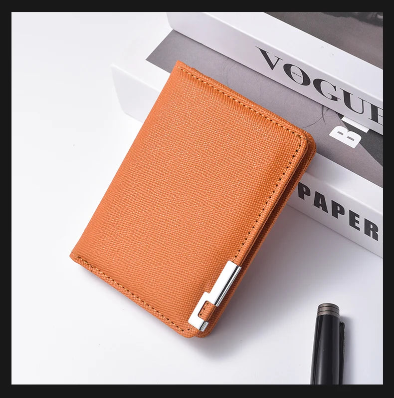 A slim vertical orange leather wallet with a silver clasp resting on a white surface next to a black pen and a magazine.