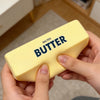 Eine Hand hält ein gelbes gesalzenes Butter-Knetspielzeug vor einem unscharfen Innenhintergrund.