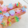 A hand displays various colorful ocean-themed resin charms including fish, sea creatures, and starfish.