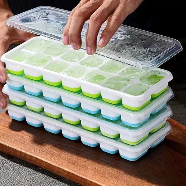 A hand is holding a stack of silicone ice cube trays filled with green ice cubes and a clear lid on a wooden surface.