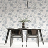 A modern dining table set with a glass top and dark chairs placed against a wall covered in a botanical leaf wallpaper.