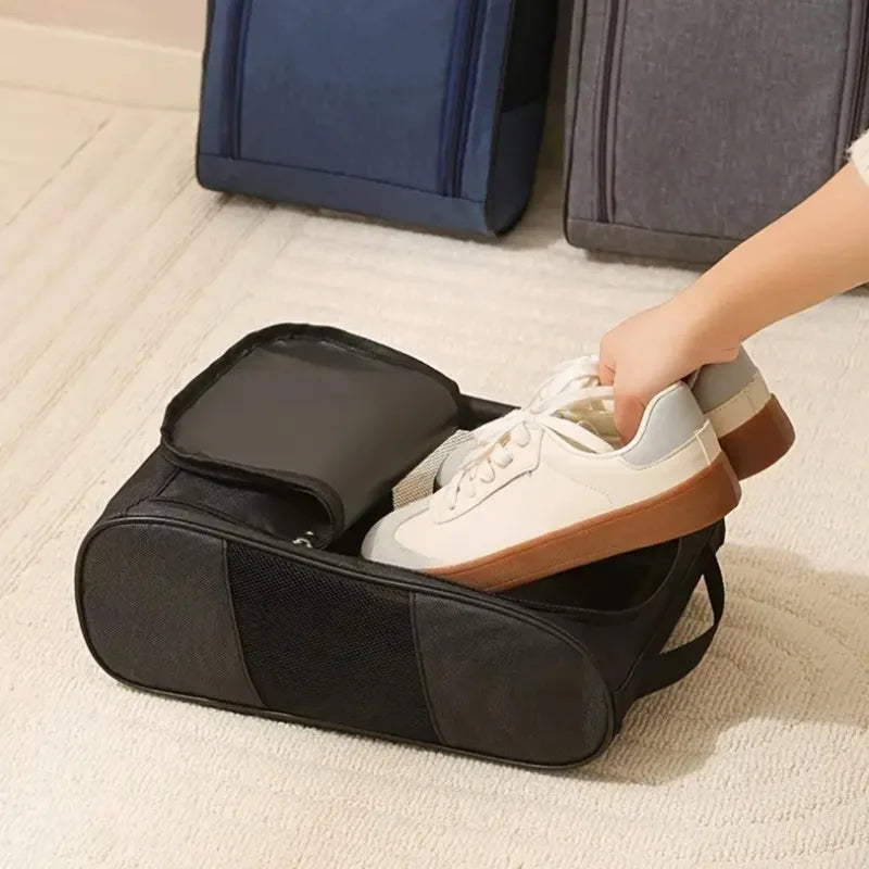 A hand is placing a pair of light-colored shoes into a black shoe storage bag on a light carpet.