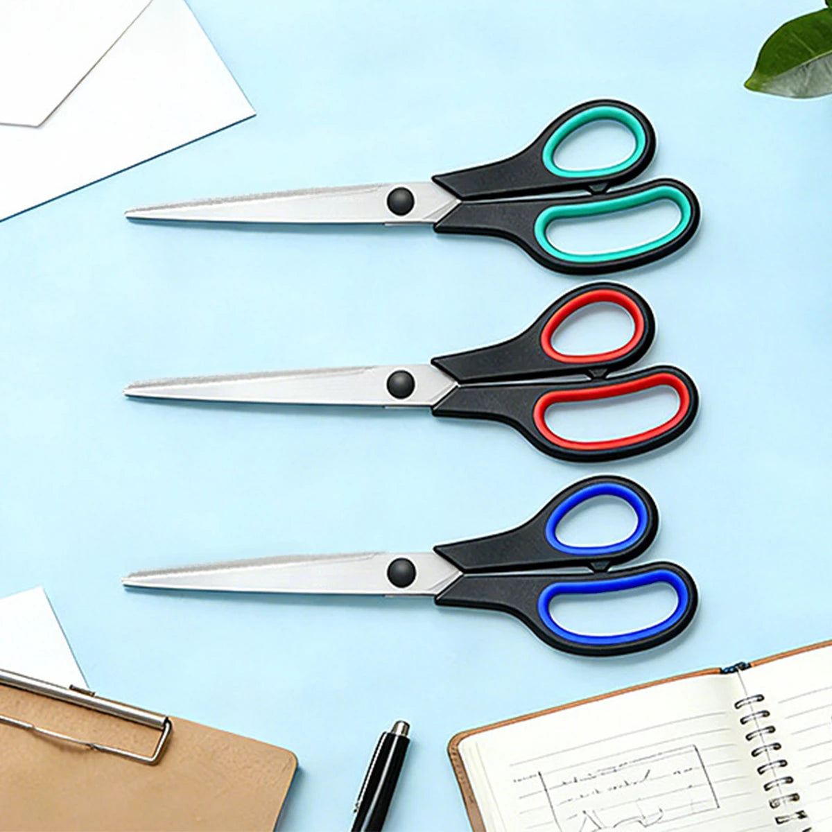 Three pairs of stainless steel scissors with soft-grip handles are displayed on a light blue background next to paper and a n