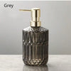 A clear glass soap dispenser with a gold pump top, set against a grey background.
