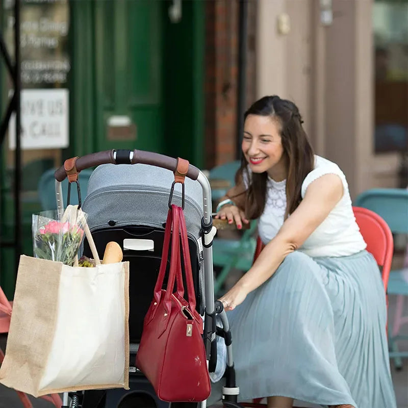 A woman sits in a stroller with a red bag and a beige tote bag, demonstrating the rotatable PU leather stroller hooks.