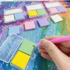 A hand is using a pink stick to apply silicone diamond painting clay pieces onto a clear plastic tray with various colored sq