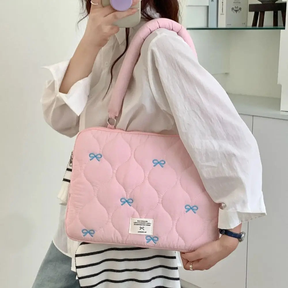 A pink quilted laptop shoulder bag with blue bows is carried by a person wearing a white shirt and striped skirt.
