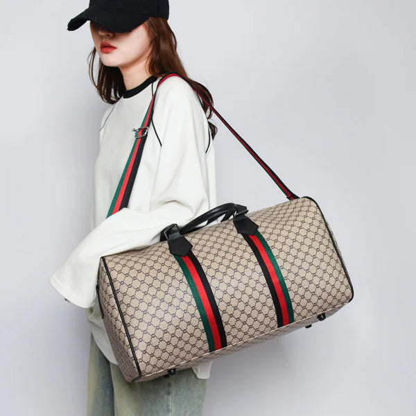 A woman holds a beige patterned travel tote bag with black and red straps against a light gray background.