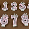 A set of rhinestone crown number candles from 0 to 9 in pink and white on a brown surface.