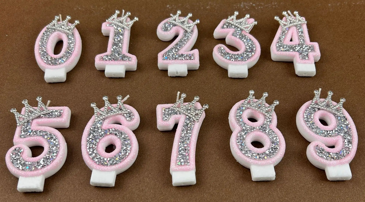 A set of rhinestone crown number candles from 0 to 9 in pink and white on a brown surface.