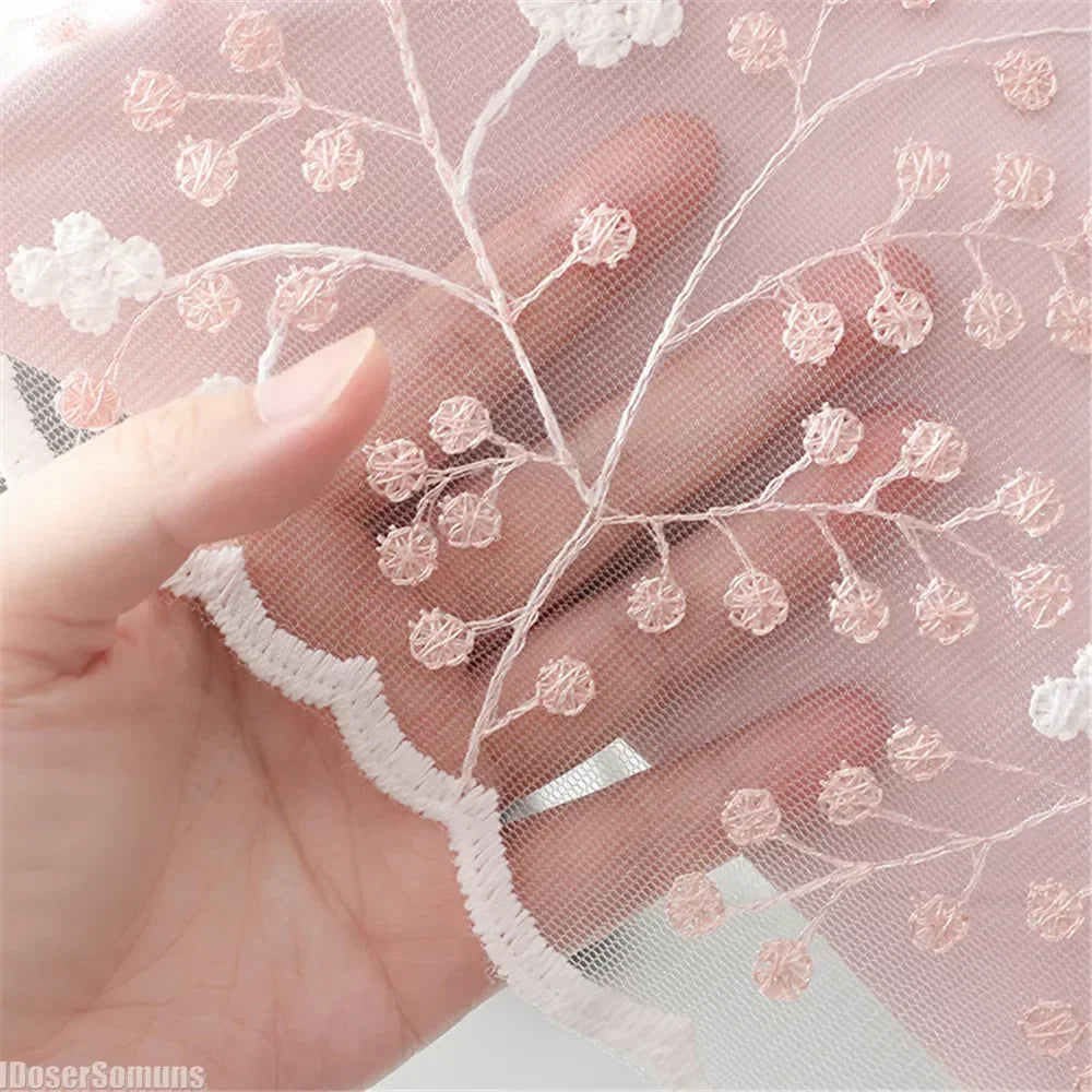 A hand holds sheer pink lace fabric featuring delicate floral embroidery and netting.