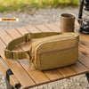 A tan tactical MOLLE waist pack is displayed on a wooden surface next to a metal mug.