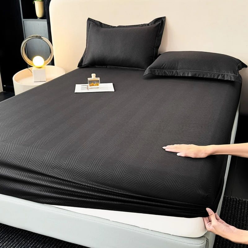 A black fitted sheet is laid out on a bed with pillows and a small bottle of lotion nearby.