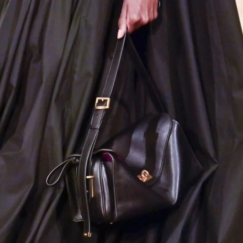 A black genuine leather square crossbody bag with a gold lock is held against a dark fabric background.