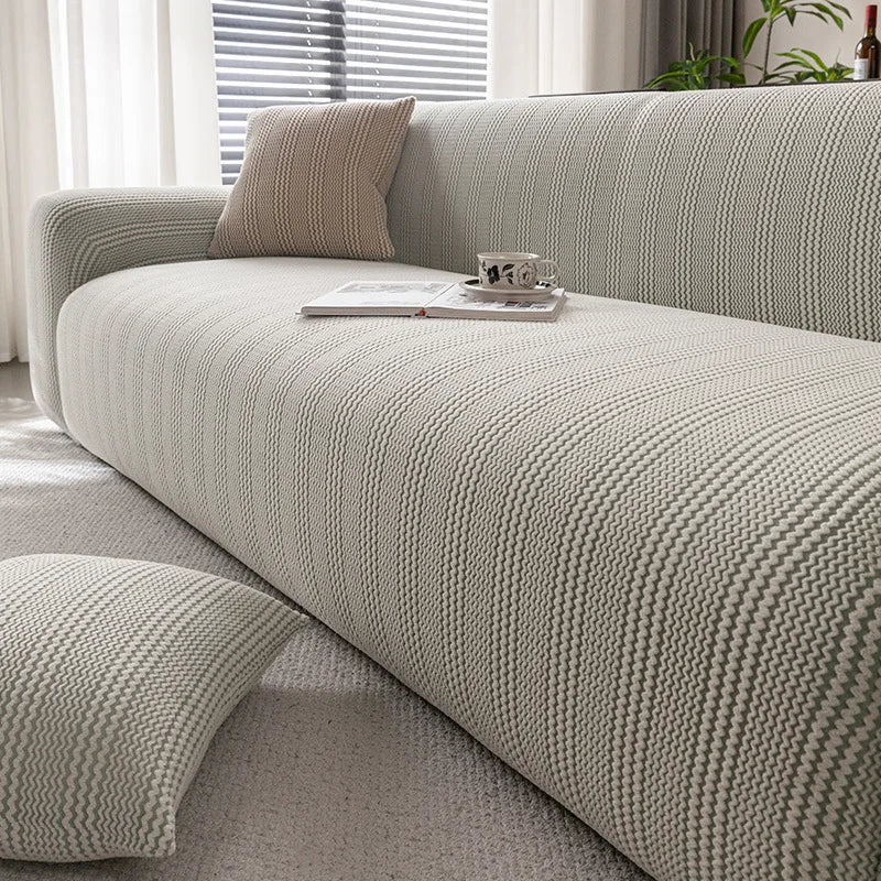 A light beige quilted stretch sofa cover with a textured weave and matching throw pillows on a modern sofa.