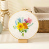 A circular embroidery hoop displays a colorful floral cross stitch design featuring yellow and purple flowers on white fabric