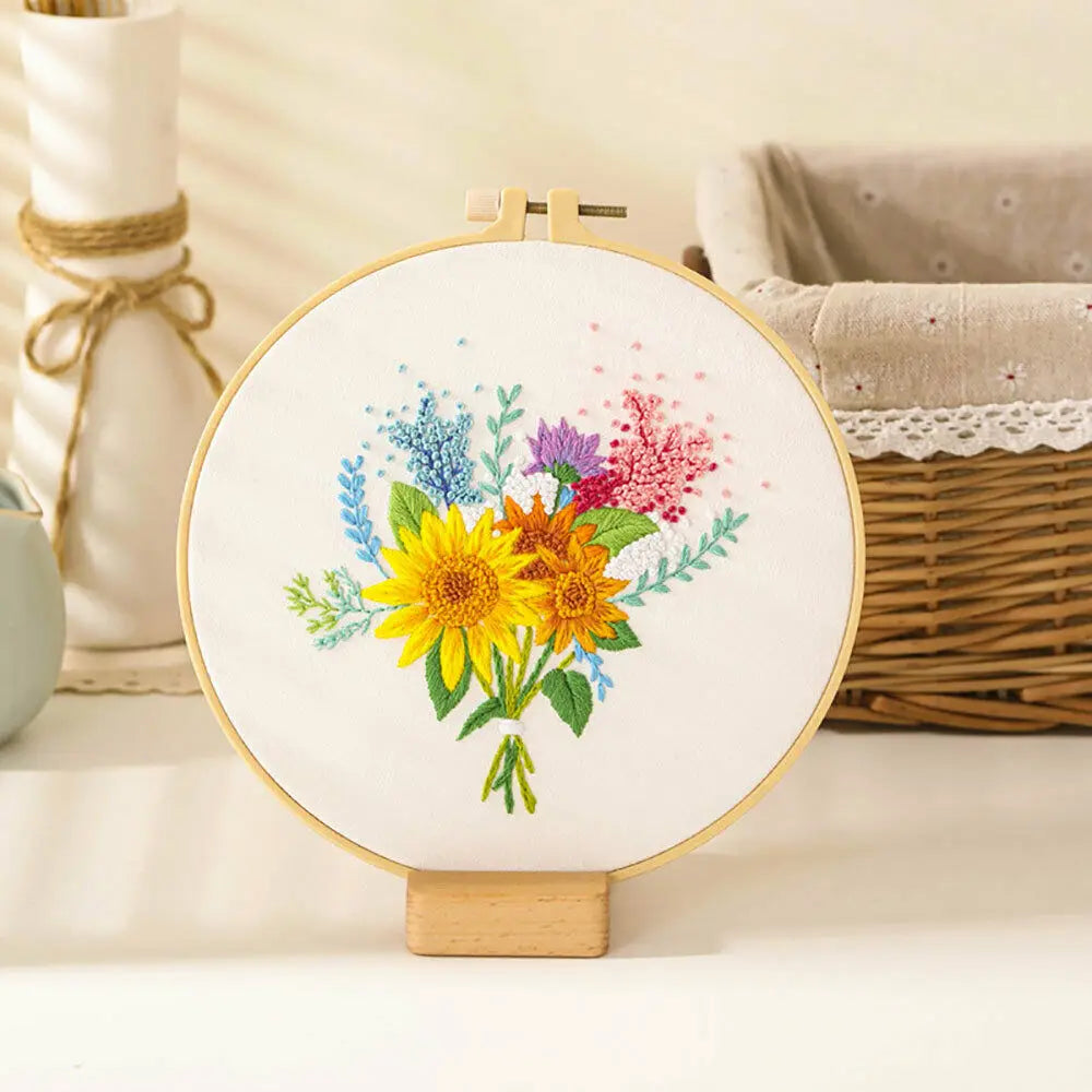 A circular embroidery hoop displays a colorful floral cross stitch design featuring yellow and purple flowers on white fabric