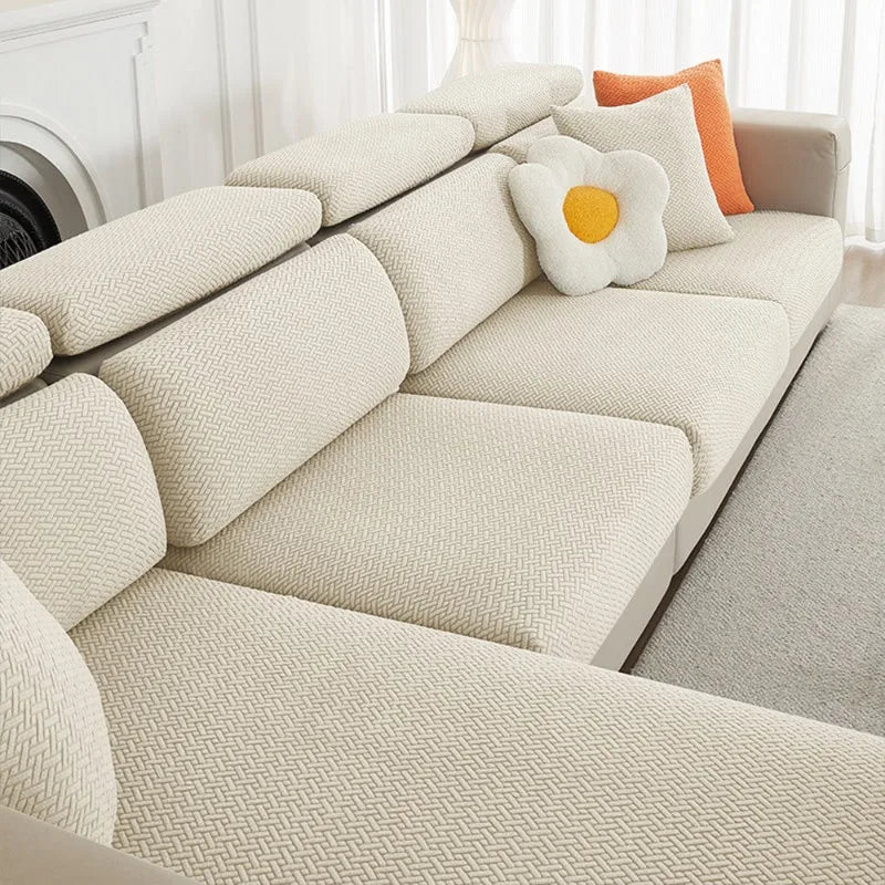 A beige jacquard elastic sofa slipcover with light yellow and orange accent pillows on a white couch.