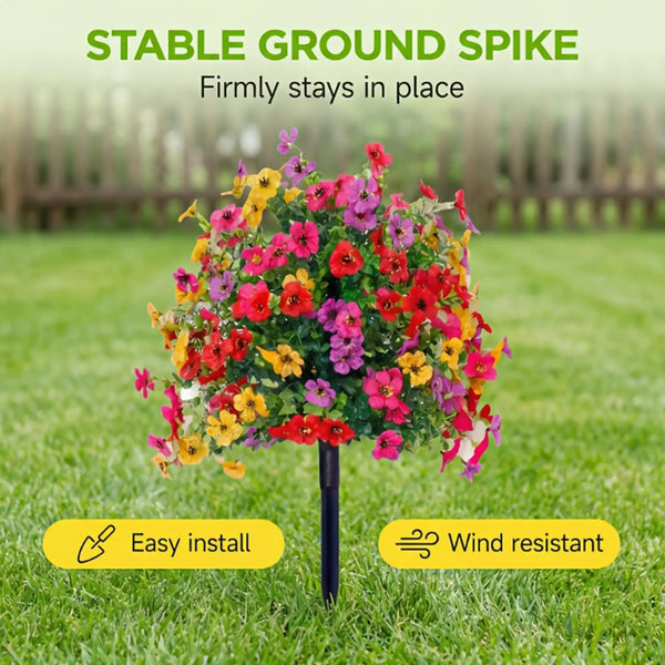 A vibrant mixed bouquet of artificial flowers in red, yellow, and pink is displayed in a black ground spike on green grass.