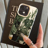A hand holds a black phone case featuring a green monstera plant pattern and a clear back panel.
