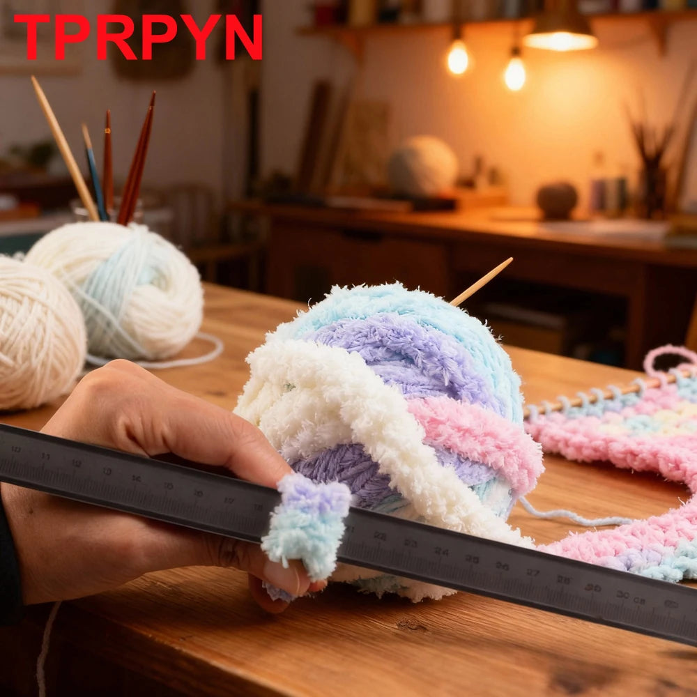 A hand measures a ball of fluffy, multi-colored chunky chenille yarn on a wooden table.