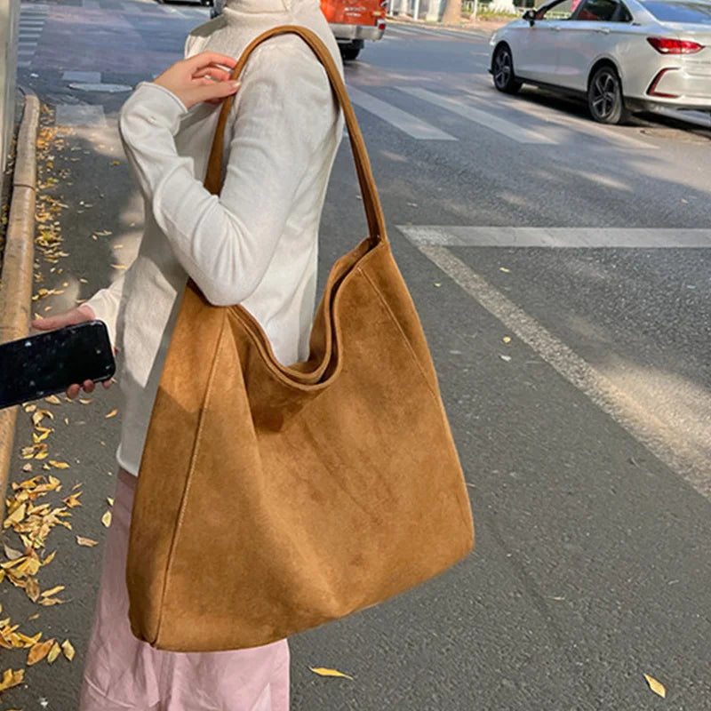 A person is holding a tan suede tote shoulder bag while standing on a street with trees and a car in the background.