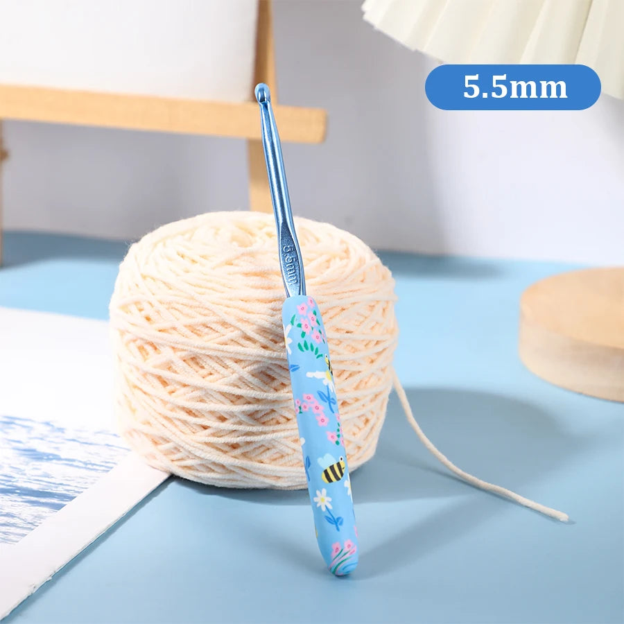A blue ergonomic crochet hook with a bee design is shown next to a ball of light beige yarn.