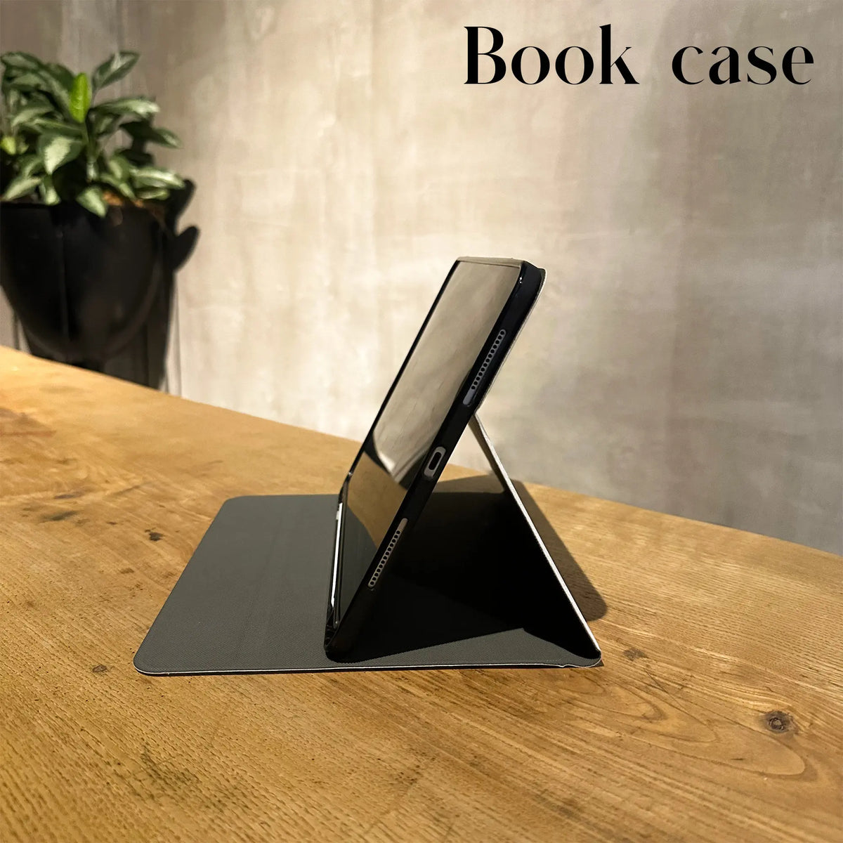 A black stand for an iPad is placed on a wooden table next to a notebook and plant.