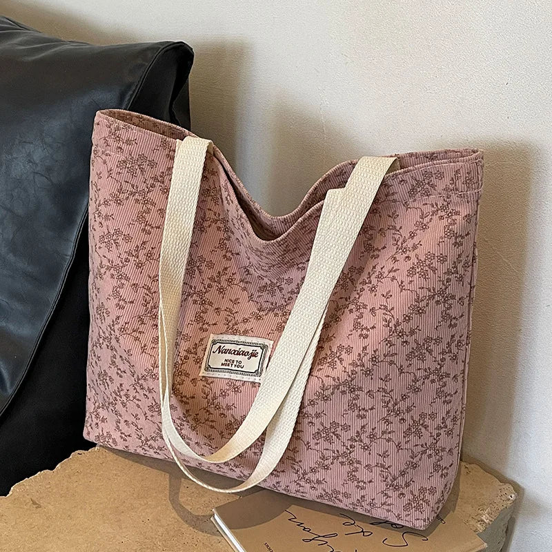 A pink corduroy tote bag with floral accents and light beige straps resting on a wooden surface.