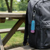 A dark gray backpack with a blue spiky sensory fidget roller attached to the zipper pull, set outdoors on a wooden bench.