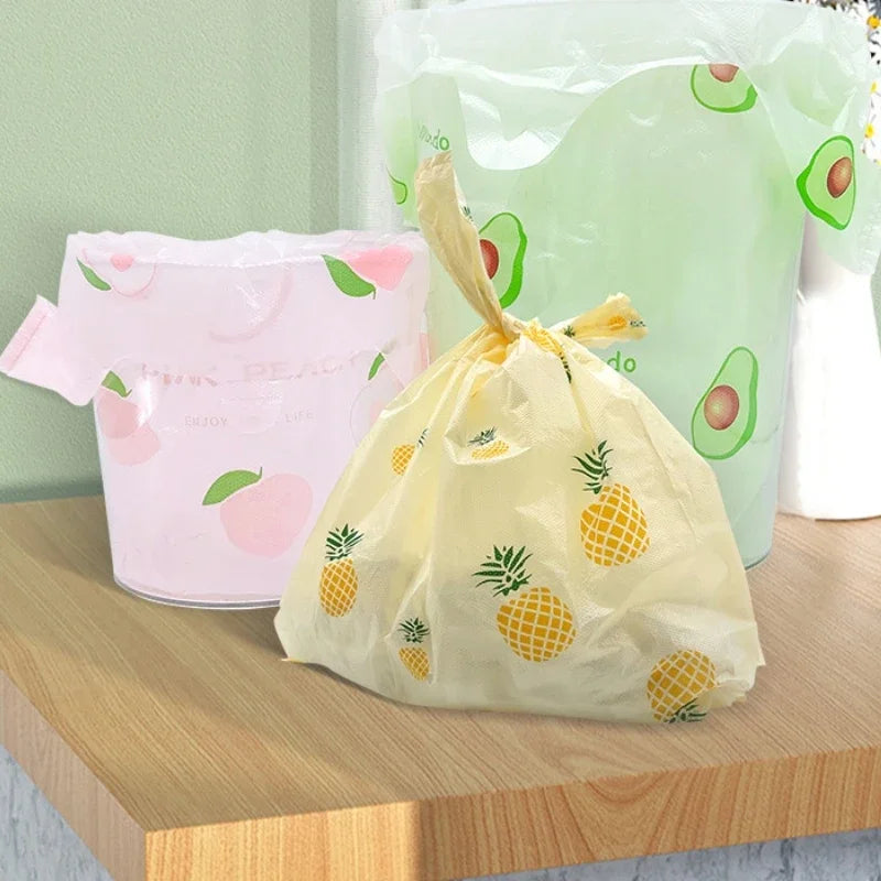 A yellow fruit-print tote bag with handles is displayed next to a large green bag and a pink bag on a wooden surface.