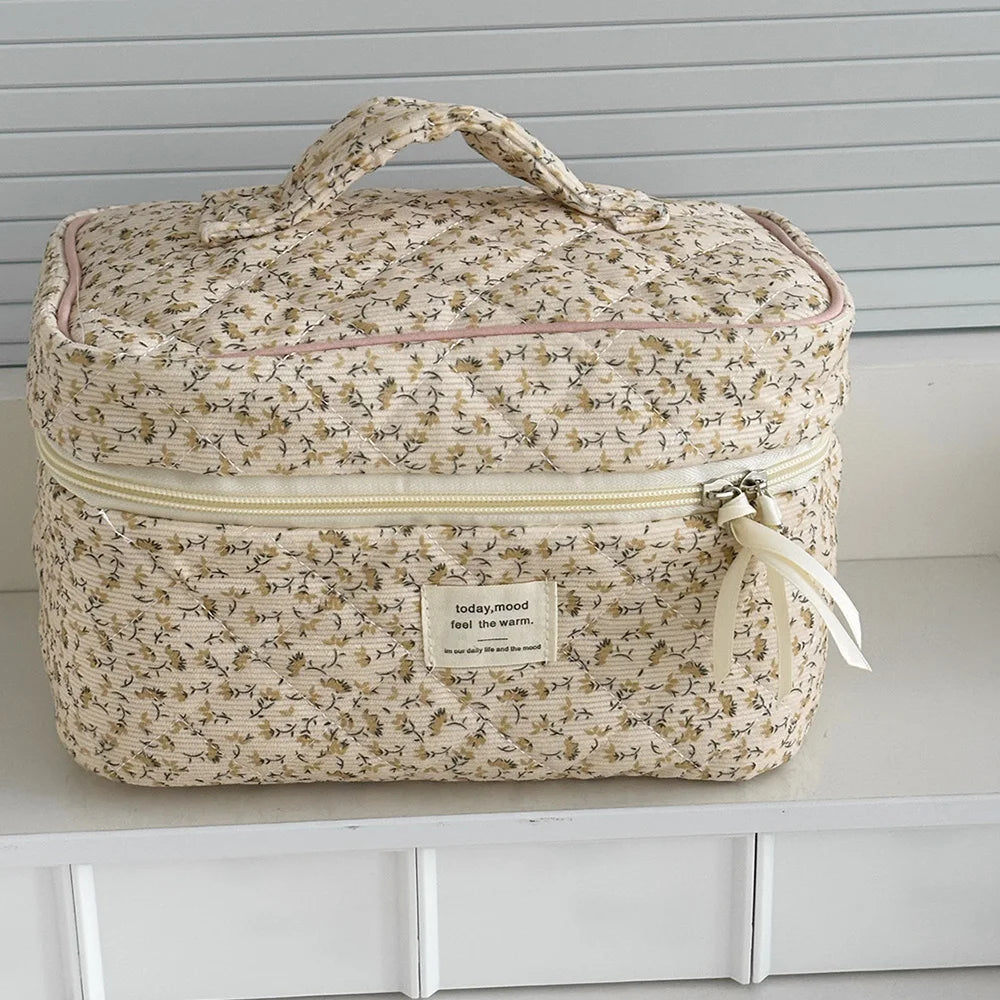 A beige floral quilted cosmetic bag with a light yellow zipper and fabric ribbon is displayed on a white shelf.