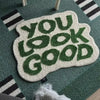 A soft, white letter rug with green embroidered text that reads YOU LOOK GOOD is placed on a teal rug.