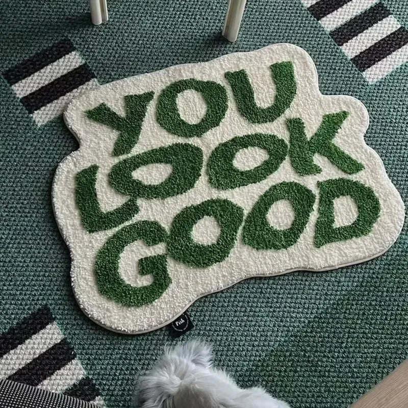 A soft, white letter rug with green embroidered text that reads YOU LOOK GOOD is placed on a teal rug.