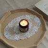 A dark jar candle sits on an oval lace embroidered placemat placed on a neutral colored plate.