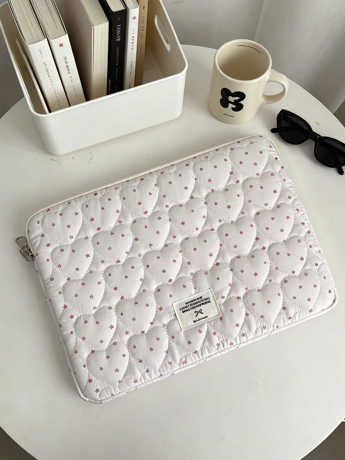 A quilted white laptop sleeve with a soft lining rests on a white table next to a mug and a box of books.