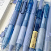 A pack of seven minimalist clear gel pens in various shades of blue and purple are displayed in a clear plastic case.