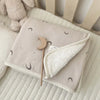A light beige floral muslin baby blanket with crescent moon patterns and a wooden teething ring is shown on a white surface.