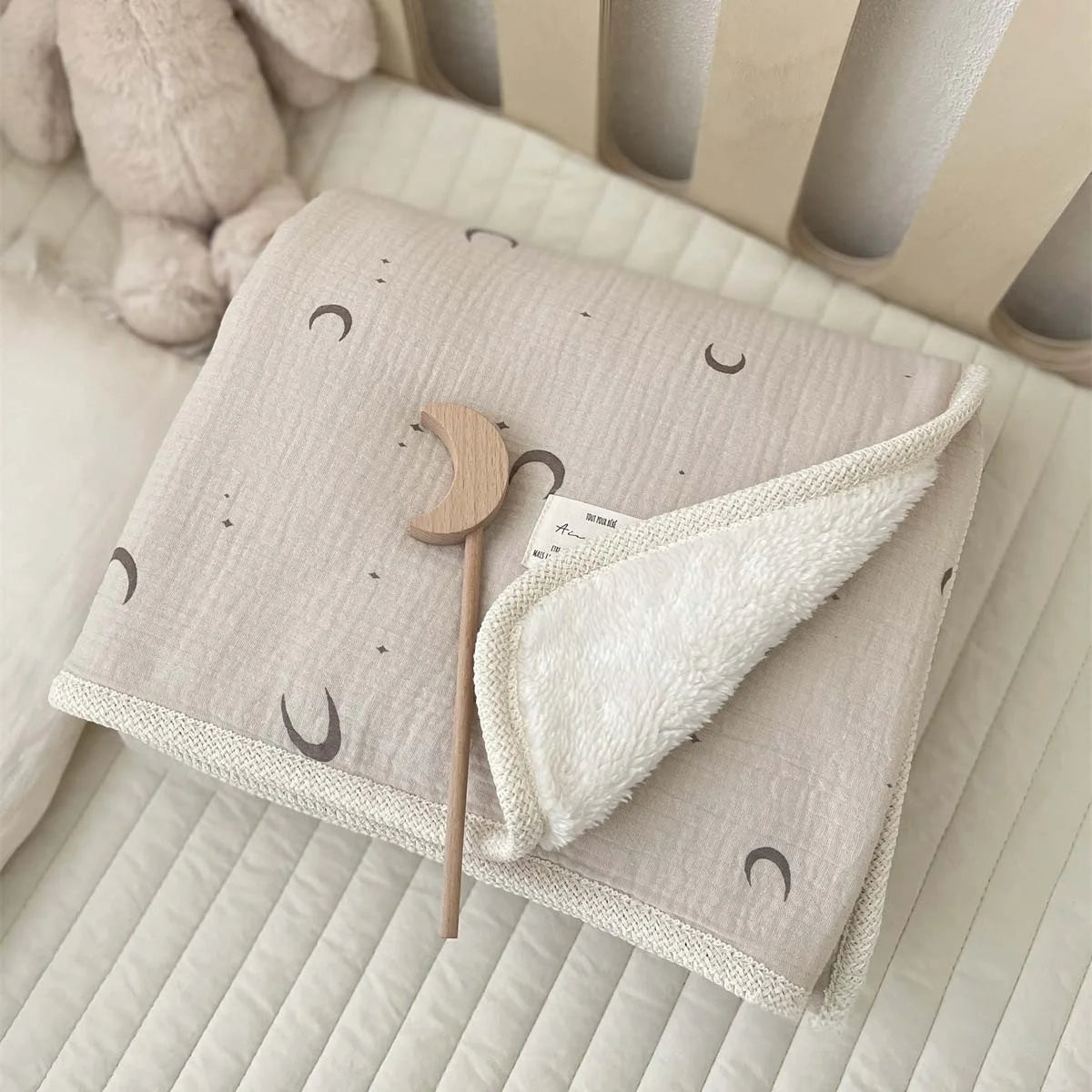 A light beige floral muslin baby blanket with crescent moon patterns and a wooden teething ring is shown on a white surface.