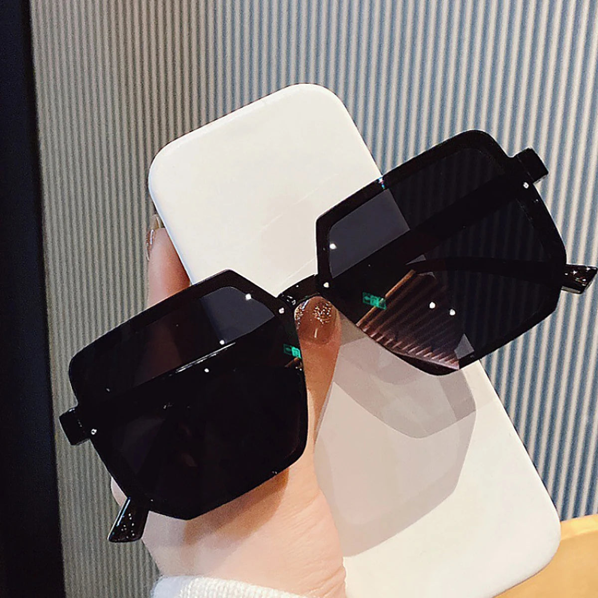 Una mano sostiene unas gafas de sol cuadradas negras vintage contra un fondo blanco.