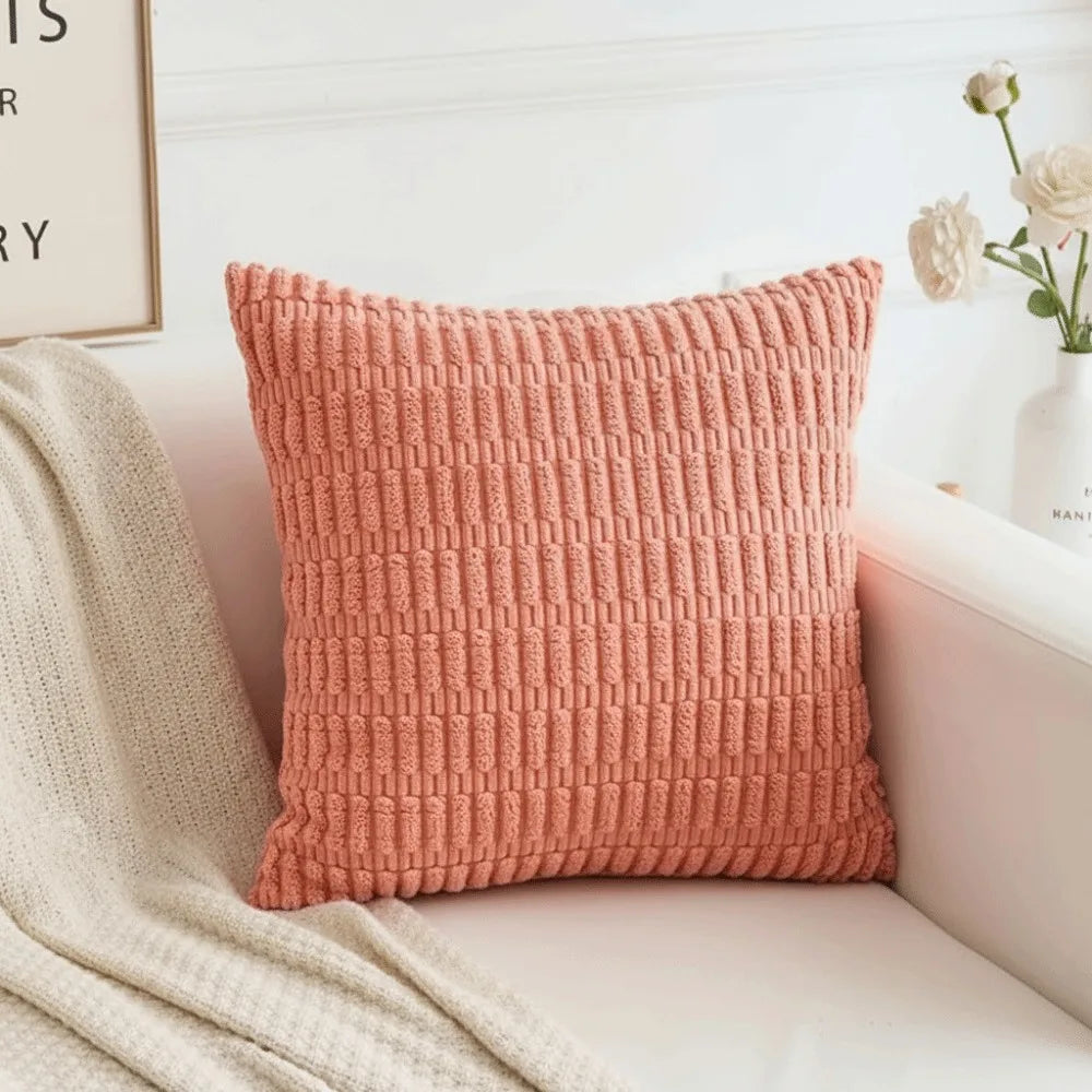 A square, textured coral pink corduroy pillow with a woven striped velvet design resting on a white surface.