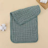 A stack of soft, sage green muslin baby burp cloths with a quilted texture and white cotton balls.